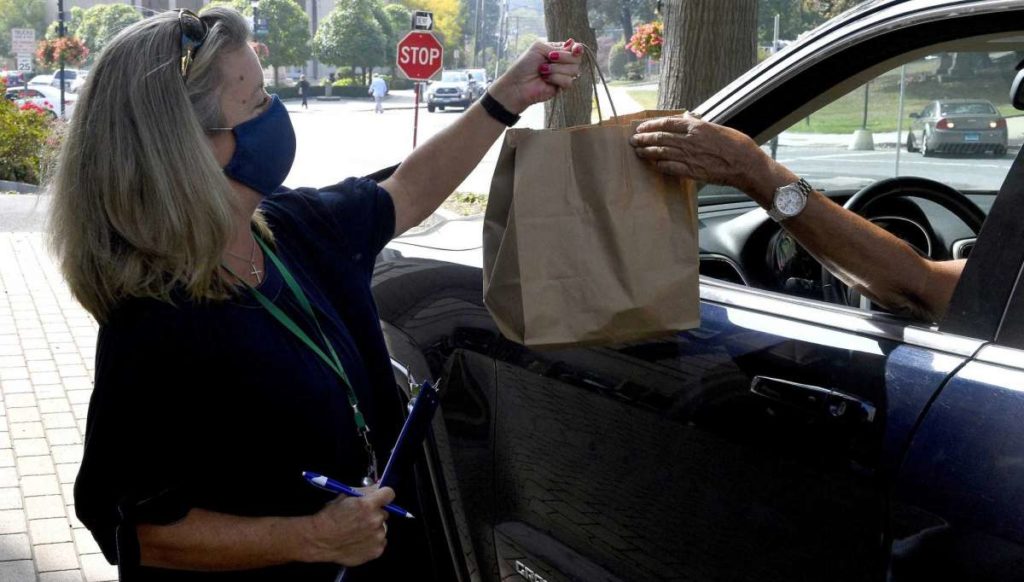 person from our senior nutrition program wearing a mask standing on a sidewalk with a clipboard and pen, handing a bag of food to an elderly person in a car