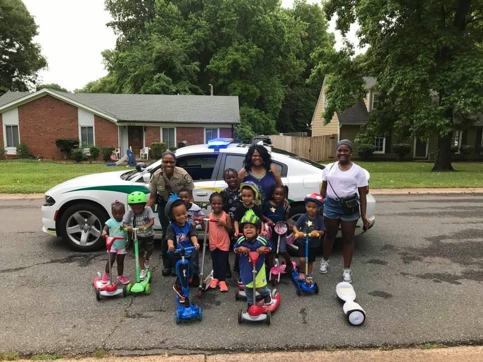 Children on scooters and their parents stand in the street with a community engagement officer in front of a Mecklenburg County Sheriff's Office car during National Night Out.