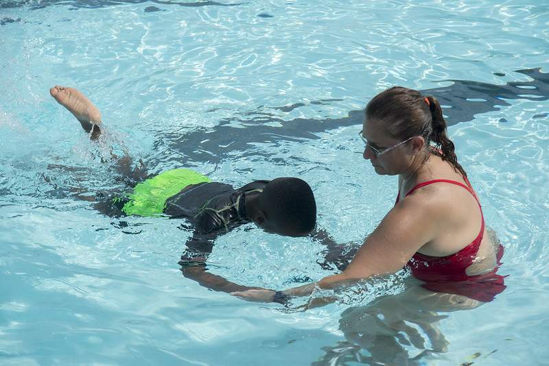 adult in red swimsuit helping a child in green swim shorts and black shirt practicing swimming 