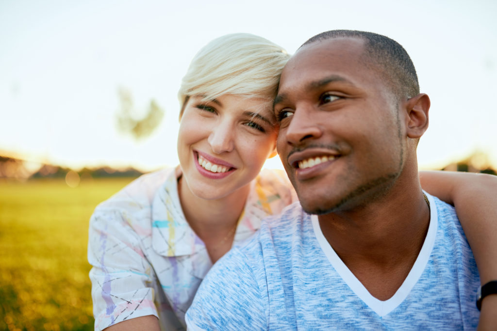 Diverse couple outside. Person on the left is staring into camera while wrapping their arm around their partner who is looking away from camera