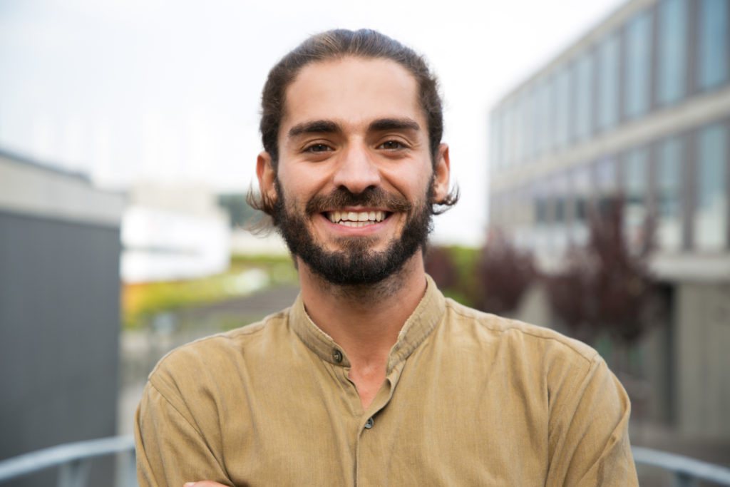 portrait of a smiling, bearded young adult standing outside of a building
