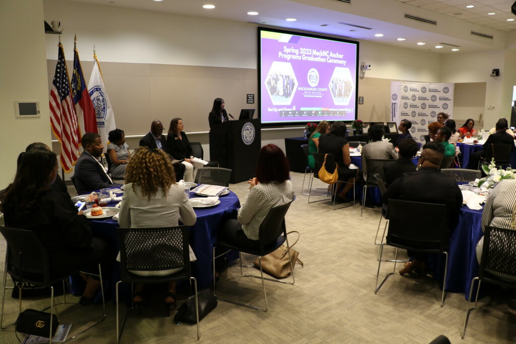 People sitting around tables in a room as a woman stands behind a podium. A screen reads Spring 2023 MeckNC Actor Programs Graduation Ceremony