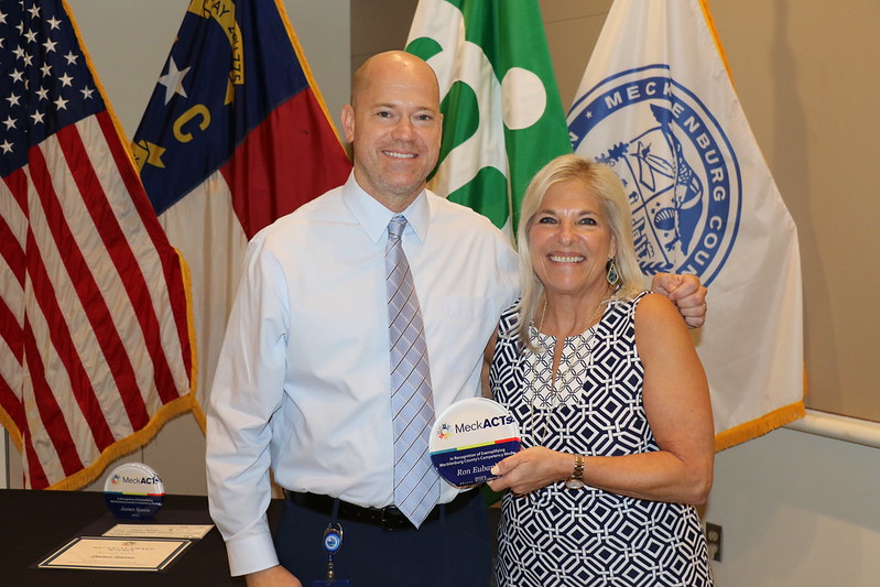 Ron Eubanks with his award and County Manager Dena R. Diorio in front of US, NC, City of Charlotte and Mecklenburg County Flags