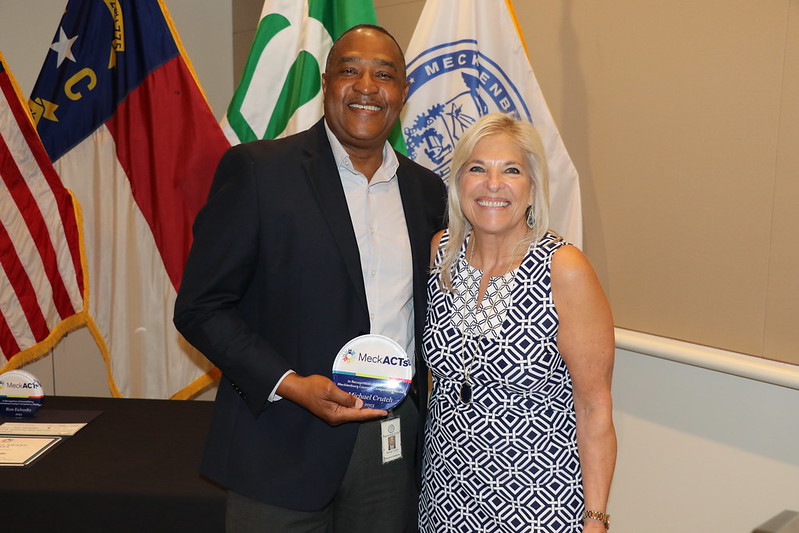 Michael Crutch with his award and County Manager Dena R. Diorio in front of US, NC, City of Charlotte and Mecklenburg County Flags