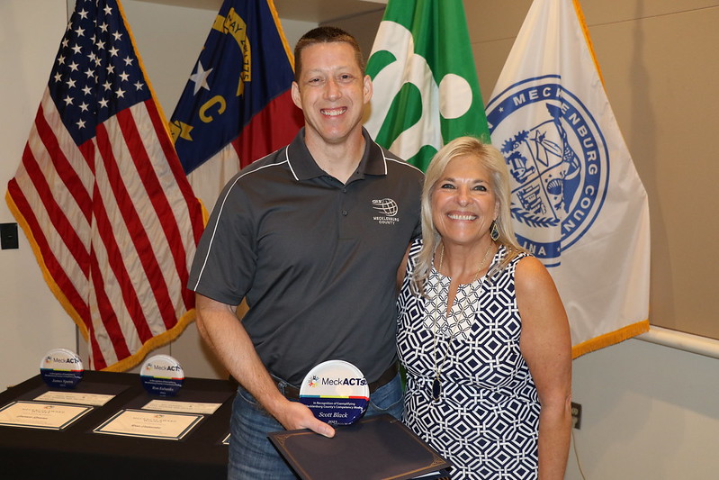 Scott Black with his award and County Manager Dena R. Diorio in front of US, NC, City of Charlotte and Mecklenburg County Flags