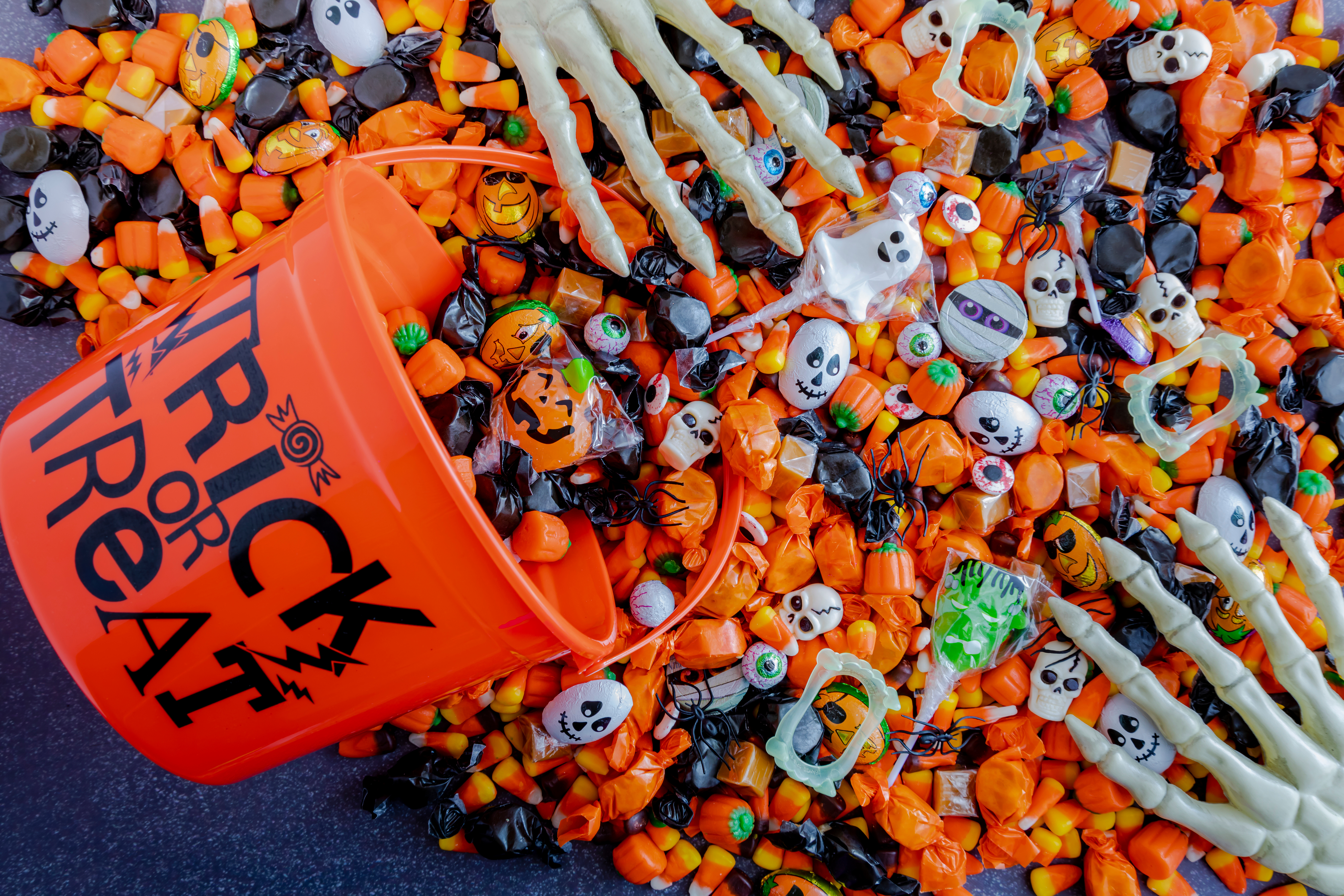 Orange trick or treat pail spilling Halloween candy on black stone surface with two skeleton hands reaching for the candy