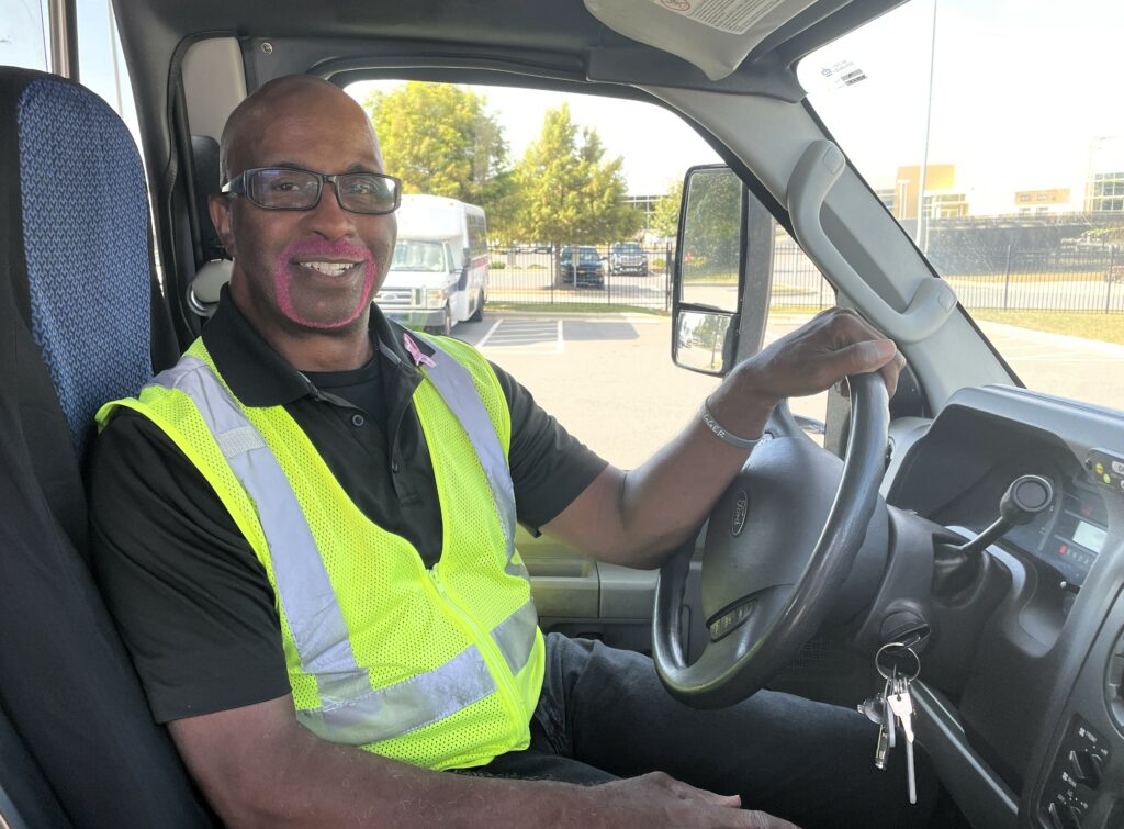 Mecklenburg Transportation Service driver J.P. Rodgers, with a pink goatee, smiling, sitting in the driver's seat of a van.