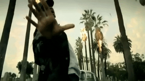 Usher dancing in front of a white car as palm trees burn behind him