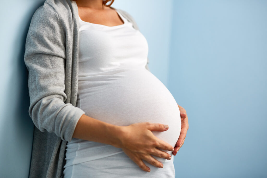 Mid-section portrait of a woman in last months of pregnancy holding her belly, standing against wall in blue room