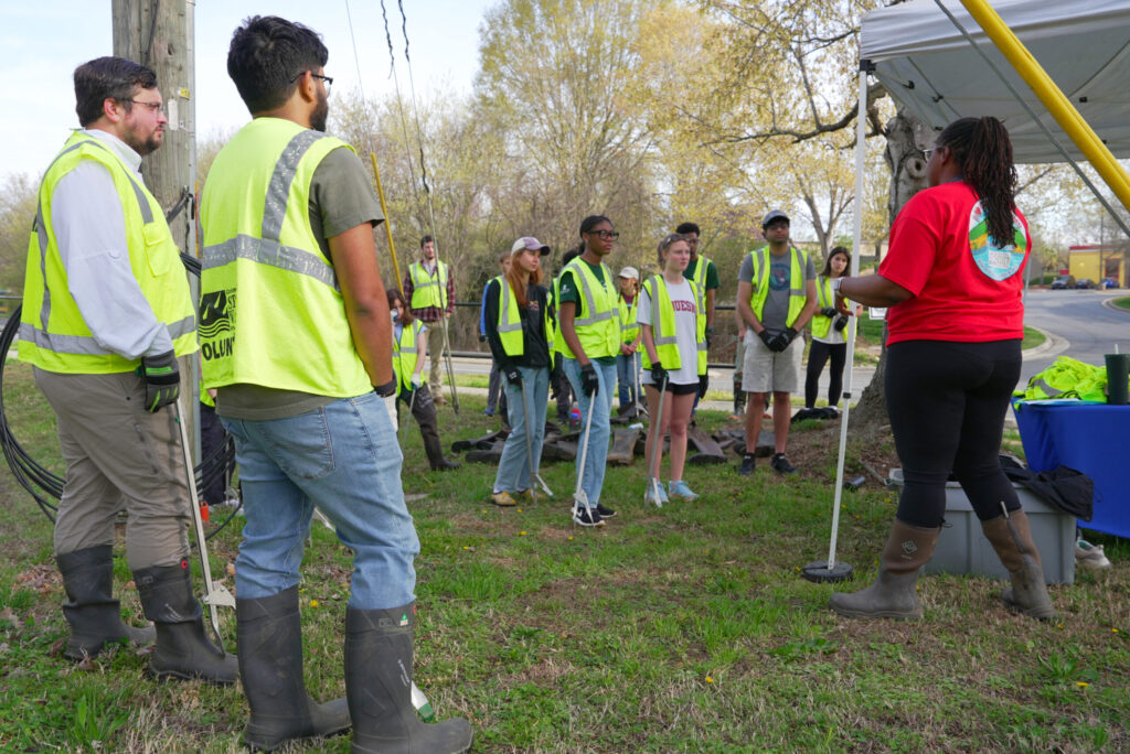 People in bright yellow safety vests with sticks to collect trash surrounded by a white tent listening to the leader