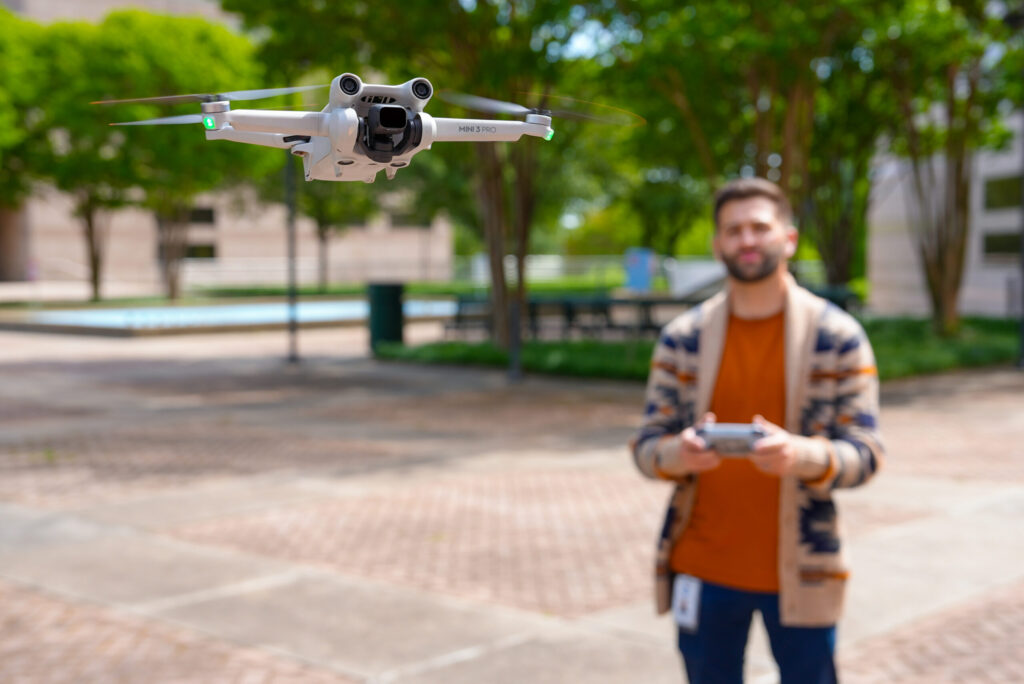 In the foreground is a drone in the air. In the background is a man holding a remote. 